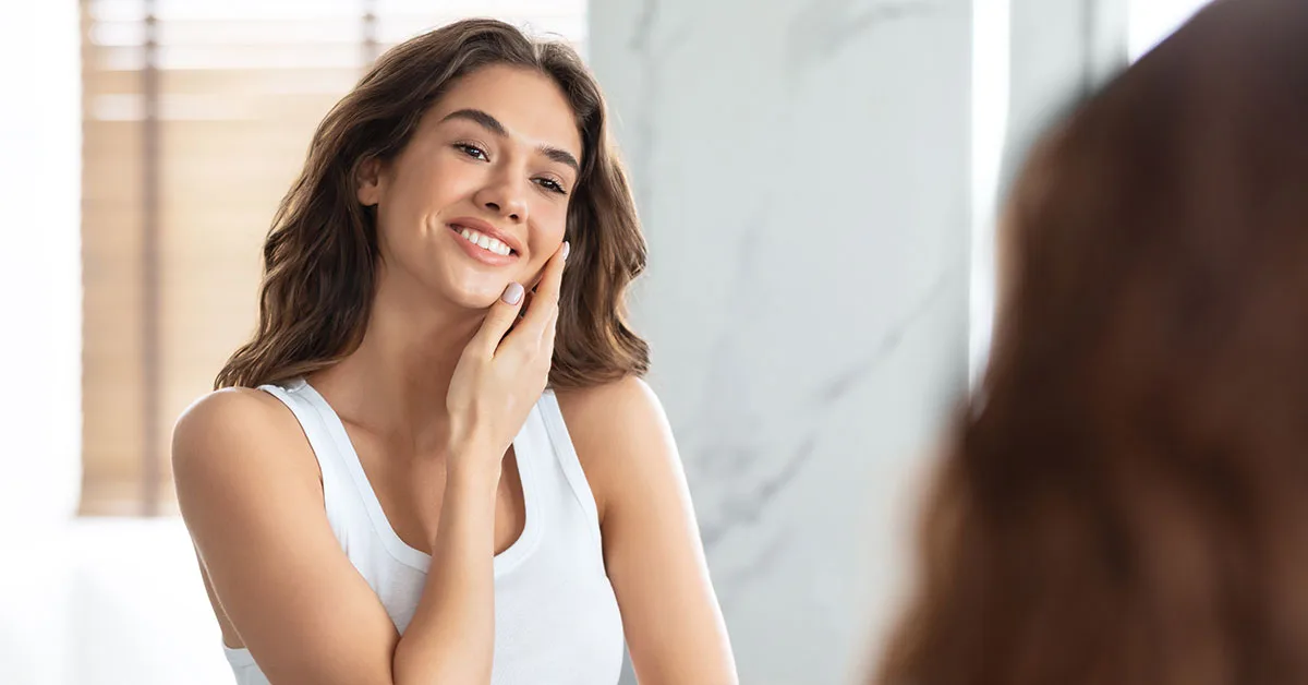 Superbe jeune femme touchant le visage soignant la peau du visage debout près d'un miroir dans une salle de bain moderne à la maison