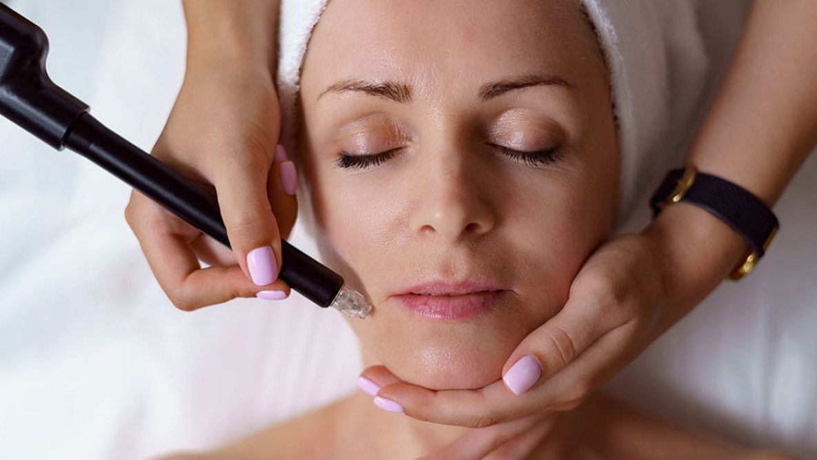 Vue du docteur cosmétologue effectuant une procédure anti-âge dans le bureau de cosmétologie. Femme satisfaite au chapeau jetable allongée sur le canapé et se relaxant