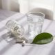 Petit pot en verre renversé laissant échapper quelques gélules blanches sur un plan de marbre clair, près d'un verre d'eau et d'une feuille verte, lumière douce de fenêtre.