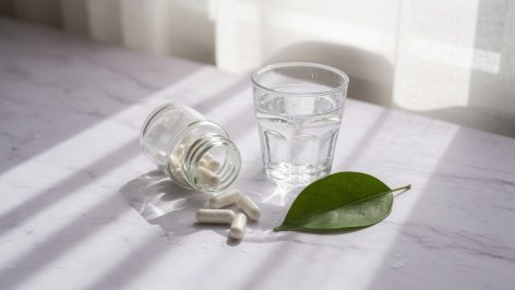 Petit pot en verre renversé laissant échapper quelques gélules blanches sur un plan de marbre clair, près d'un verre d'eau et d'une feuille verte, lumière douce de fenêtre.